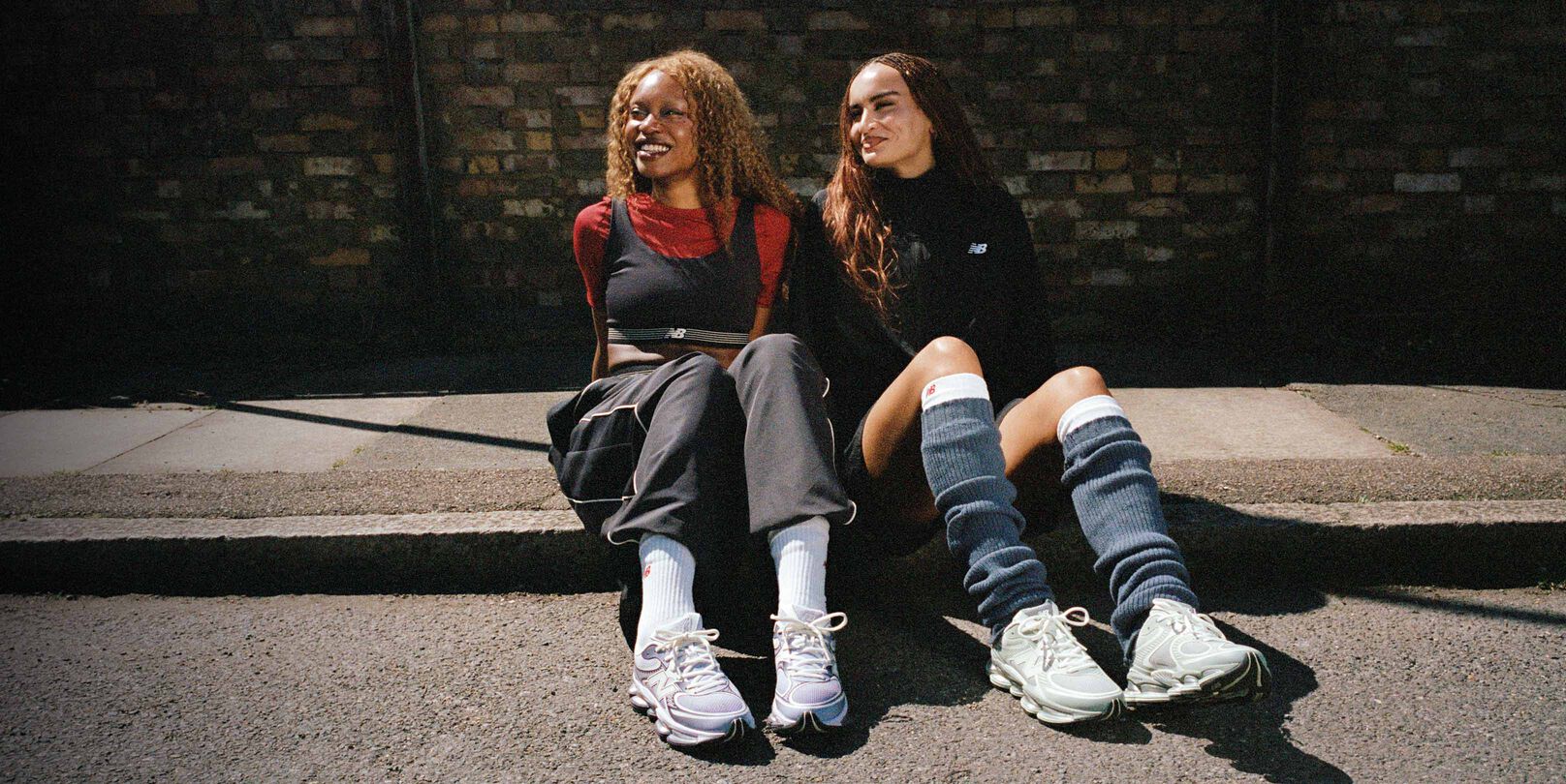   Two women seated on a sidewalk wearing New Balance sneakers.